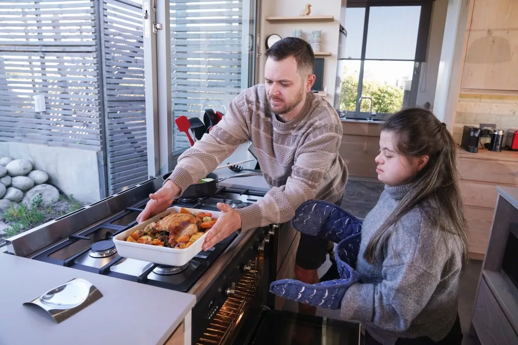 Support worker helping disabled girl with down syndrome make roast chicken at home